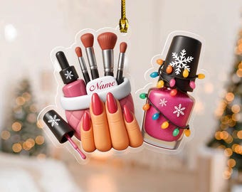 May include: A Christmas ornament featuring nail polish, makeup brushes, and a hand with red nails. A bottle of pink nail polish is decorated with a string of colorful lights and a snowflake. The ornament has a gold string for hanging.