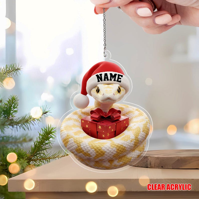 May include: A clear acrylic Christmas ornament depicting a yellow and white snake wearing a Santa hat with the word "NAME". The snake is coiled around a red gift box with a red bow. The words "CLEAR ACRYLIC" are at the bottom.