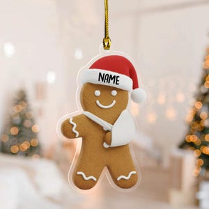 May include: A gingerbread man Christmas ornament wearing a Santa hat with the word "NAME" and a white arm sling. The ornament is brown with white icing details and a gold string for hanging.