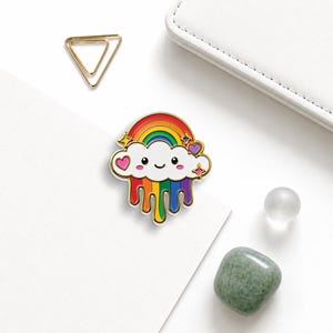 May include: A gold-toned enamel pin featuring a white cloud with a smiling face, topped with a colorful rainbow arch and dripping rainbow-colored paint from the bottom. The design includes small pink hearts and four-pointed star accents in gold.