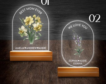 May include: Two illuminated acrylic signs with floral designs and personalized messages. The first sign reads "Best Mom Ever" with yellow daffodils and the names Amelia, Andrew, and Andie. The second sign reads "We Love You" with purple flowers and the names Sophia, Addie, and Liliana.