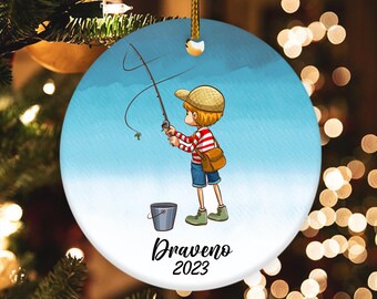 May include: A blue and white Christmas ornament featuring a cartoon boy fishing with a red and white striped shirt, a brown bucket, and the text "Draveno 2023".