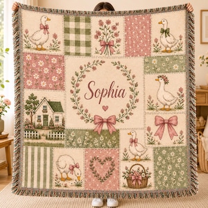 May include: A woven throw blanket featuring a patchwork design with illustrations of ducks, a house, flowers, bows, and a heart. The center panel displays the name Sophia inside a floral wreath. The blanket has a fringed edge and muted tones.