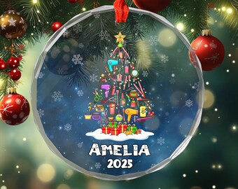 May include: A clear, round Christmas ornament with a red ribbon. Inside, a Christmas tree is formed from salon tools, with the name "AMELIA" and the year "2025" at the bottom. The background is a snowy blue.