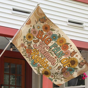 May include: A beige flag with the text "BIG FAN OF Human Rights" in orange, pink, and teal. The flag is decorated with colorful flowers and butterflies. The flag is hanging outside a house.