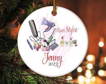 May include: A white ceramic Christmas ornament featuring a hair stylist theme. The ornament has illustrations of hair styling tools, makeup, and the text "Hair Stylist" and "Jenny 2023" in red script. A gold string is attached for hanging.