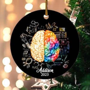 May include: A black round ornament with a gold string for hanging. The ornament features a watercolor illustration of a brain, split into two halves. One half is beige, the other is a rainbow of colors. The name "Addison" and the year "2023" are printed on the ornament.