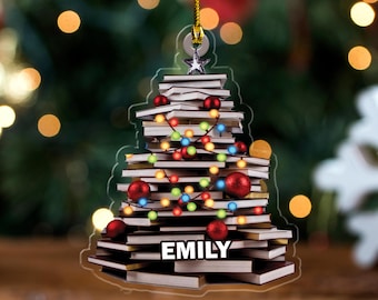 May include: A Christmas tree ornament shaped like a stack of books, decorated with red ornaments and colorful lights. The ornament has the name "EMILY" printed on it and is suspended by a gold string.