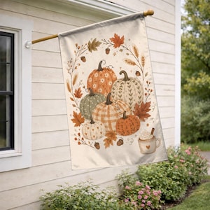 May include: A rectangular garden flag featuring a cream-colored background with a central illustration of five pumpkins in shades of orange, green, and white, surrounded by autumn leaves, wheat stalks, acorns, and a mug of hot cocoa with a heart.