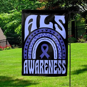 May include: Black garden flag with the words "ALS AWARENESS" in light blue, above a light blue rainbow design and a purple awareness ribbon. The flag is hanging in a garden.