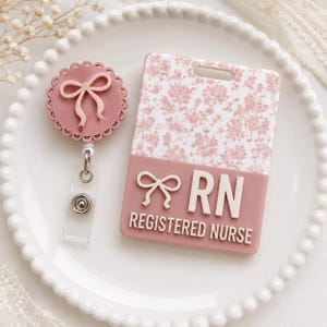 May include: A dusty rose badge reel with a scalloped edge and a raised bow, paired with a matching rectangular ID badge holder featuring a white and dusty rose floral pattern, the letters RN, the text REGISTERED NURSE, and a raised bow accent.