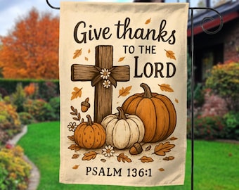 May include: A beige garden flag with the text "Give thanks to the Lord" and "Psalm 136:1". The flag features a wooden cross, pumpkins, leaves, and acorns in shades of brown, orange, and white. The flag is hanging on a black metal stand.