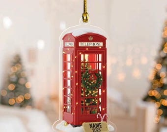 May include: A red telephone booth Christmas ornament with a wreath and lights inside. The ornament has the word "TELEPHONE" on top and a small sign that says "NAME 2025". It is hanging from a gold string.