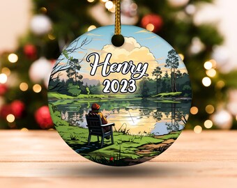 May include: Round ceramic ornament featuring a serene lake scene with a person fishing from a chair. The ornament has the name "Henry" and the year "2023" in a decorative font. The background shows trees, water, and a sunset.