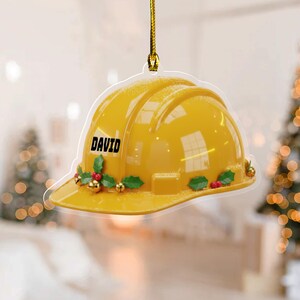 May include: A yellow construction helmet ornament with the name "DAVID" in black lettering. The ornament is decorated with holly leaves, red berries, and gold accents. It hangs from a gold string, perfect for holiday decor.