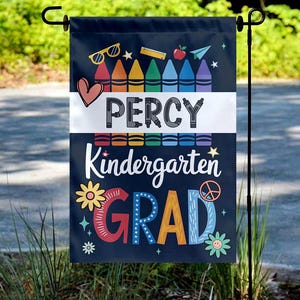 May include: A navy blue garden flag with the name "Percy" and the words "Kindergarten GRAD" in colorful letters. The flag features crayon illustrations, a heart, peace sign, and other school-themed graphics. The flag is hanging on a black metal stand.