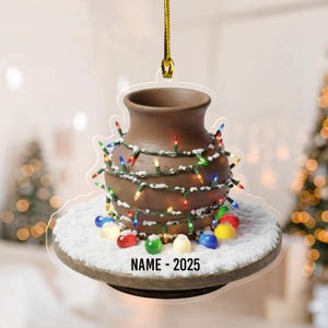 May include: A brown ceramic vase Christmas ornament, adorned with colorful string lights and faux snow. The ornament has a gold string for hanging and the text "NAME - 2025" at the bottom. Festive holiday decor.