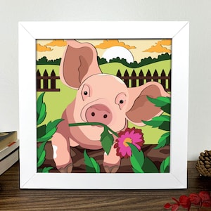 May include: A pink piglet with large ears is standing in a field of green grass and flowers. The piglet is holding a pink flower in its mouth. There is a wooden fence behind the piglet.