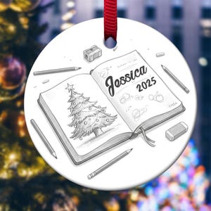 May include: A white, round Christmas ornament with a red ribbon. The ornament features a pencil sketch of a Christmas tree and a notebook with the name "Jessica" and the year "2025". Pencils, a pencil sharpener, and an eraser surround the notebook.