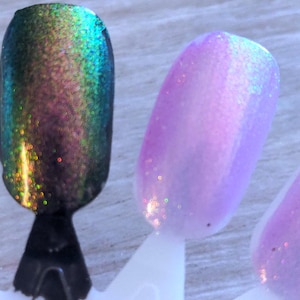 May include: Close-up of nail polish swatches. One swatch is a dark green with iridescent shimmer, transitioning to blue and purple. Another is a light purple with a subtle glitter effect. The swatches are on white plastic sticks.