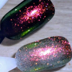 May include: Two nail polish samples showcasing a glitter effect. The top sample is a dark green with pink glitter, while the bottom sample is a lighter green with pink glitter. Both samples are on white sticks.