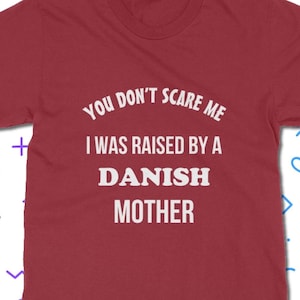 Może przedstawiać: Bordowa koszulka z białym napisem: "YOU DON'T SCARE ME I WAS RAISED BY A DANISH MOTHER". Koszulka ma okrągły dekolt i kr&oacute;tkie rękawy. Projekt jest wyśrodkowany z przodu.