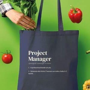 May include: A navy blue tote bag featuring white text that reads Project Manager, a professional herder of cats, and someone who thinks 9 women can make a baby in 1 month. The bag is surrounded by floating fresh vegetables including celery and peppers.