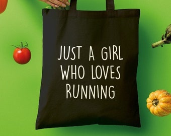 Puede incluir: Una bolsa de tela negra con el texto "Just a girl who loves running" en blanco. La bolsa está sostenida por una mano y rodeada de verduras y frutas, incluyendo un pimiento rojo, una verdura de hoja verde, un tomate rojo, un tomate amarillo y una pequeña calabaza amarilla.