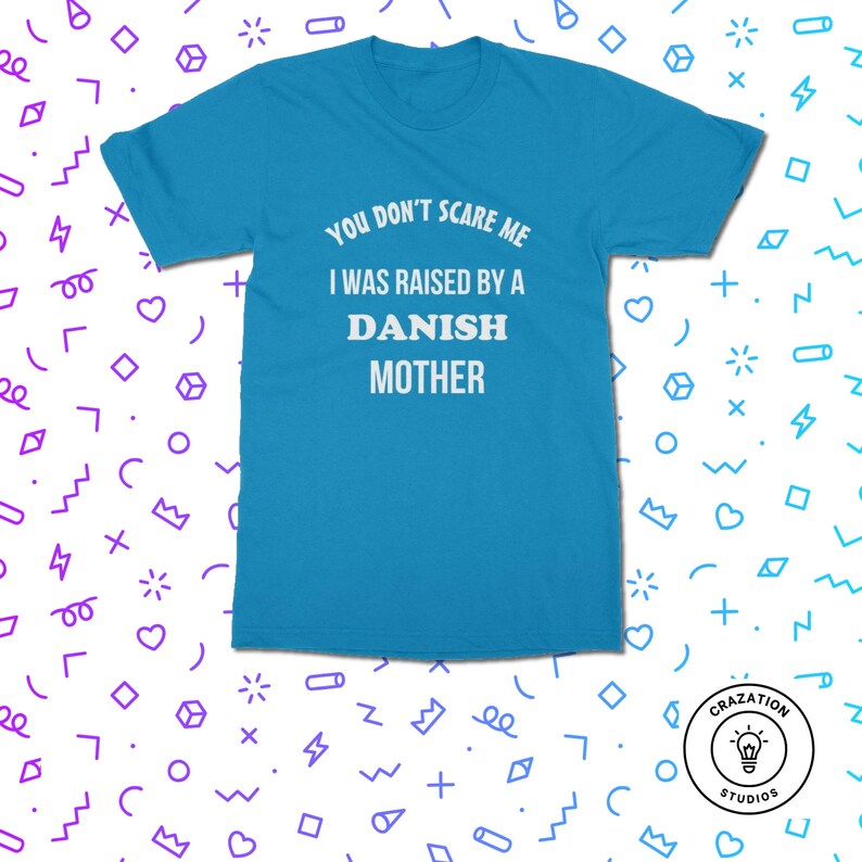 Może przedstawiać: Turkusowa koszulka z białym napisem: "YOU DON'T SCARE ME I WAS RAISED BY A DANISH MOTHER". Koszulka ma kr&oacute;tkie rękawy i okrągły dekolt. Logo Crazation Studios znajduje się w prawym dolnym rogu.