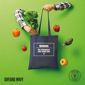 Puede incluir: Una bolsa de tela azul marino oscuro con un borde rectangular blanco y el texto WARNING MAY SPONTANEOUSLY START TALKING ABOUT AI. La bolsa está rodeada de verduras frescas flotantes, incluyendo apio, un pimiento rojo, un tomate y calabaza.