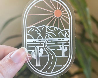 May include: Oval sticker featuring a desert landscape design. The sticker depicts a road winding through a desert scene with mountains, cacti, and a sunburst design. The color palette includes shades of brown, white, and orange.