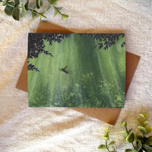 May include: A greeting card featuring a painting of a hummingbird in flight over a green forest scene. The artwork is set against a backdrop of vertical light rays, with dark tree branches at the top. The card is placed on a brown envelope.