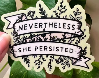 Puede incluir: Una pegatina verde claro con una pancarta blanca que dice "Nevertheless, She Persisted." La pancarta está rodeada de dibujos de flores y hojas en línea negra.
