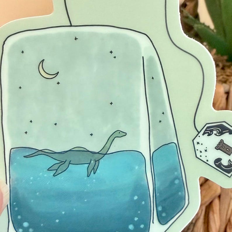 May include: A whimsical sticker featuring a light green tea bag shape. Inside, a blue lake scene with a Loch Ness Monster illustration, a crescent moon, and stars. The tea bag has a black outline.