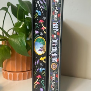 May include: Two hardcover books. One has a black spine with colorful illustrations of mushrooms, flowers, and a winter scene. The other book is dark green with gold lettering that reads "Emily Wilde's Map of the Otherlands."