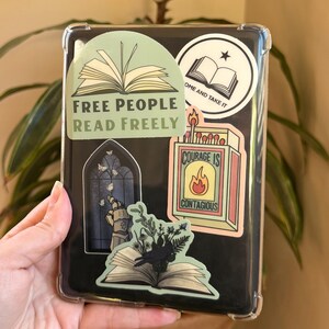 Puede incluir: Un lector de libros electr&oacute;nicos negro en una funda transparente, adornado con varias pegatinas. Las pegatinas presentan frases como "Free People Read Freely" y "Courage is Contagious", junto con ilustraciones de libros y una caja de cerillas.