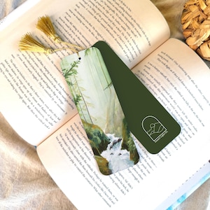 May include: A green bookmark with a landscape painting of a waterfall and trees. The bookmark has a gold tassel and the text "Desert Light Co." on the bottom right. The bookmark is placed in an open book.