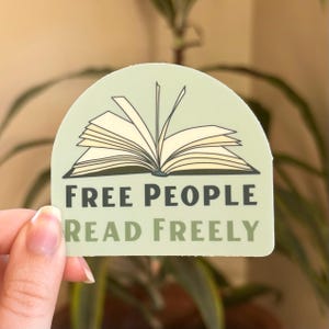 Puede incluir: Una pegatina verde claro con el texto "FREE PEOPLE READ FREELY" y una ilustraci&oacute;n de un libro abierto. La pegatina tiene un borde superior redondeado y est&aacute; sujeta por una mano frente a un fondo verde borroso.