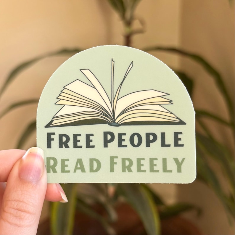 Puede incluir: Una pegatina verde claro con el texto "FREE PEOPLE READ FREELY" y una ilustraci&oacute;n de un libro abierto. La pegatina tiene un borde superior redondeado y est&aacute; sujeta por una mano frente a un fondo verde borroso.