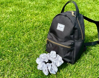 May include: A black mini backpack with gold zips and a white label. A silver scrunchie sits beside the backpack on green grass. The backpack has a top handle and adjustable straps.