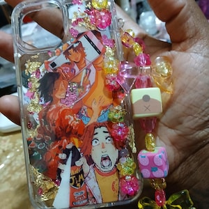 May include: Clear phone case with a colorful anime-style graphic featuring multiple characters. The case is decorated with pink and yellow beads, gold flakes, and a charm. The graphic includes the text "Trash".