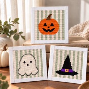 May include: Three square cross-stitch art pieces featuring Halloween motifs. One displays an orange jack-o'-lantern, another a white ghost, and the third a black witch hat with a purple band. Each is set against a light green striped background.