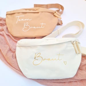 Könnte beinhalten: Zwei Bauchtaschen, eine beige und eine cremefarbene, mit verstellbaren Trägern. Die beige Tasche trägt den Schriftzug "Team Braut" in weißer Schrift. Die cremefarbene Tasche hat das Wort "Braut" in goldener Schrift mit einem kleinen Herz.