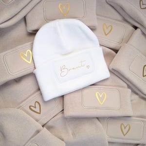 Könnte beinhalten: Eine Sammlung von beigen und weißen Strickmützen. Die weiße Mütze trägt den Schriftzug "Braut" in Gold. Beige Mützen haben ein goldenes Herzdesign. Die Mützen sind ein weiches, warmes Accessoire.