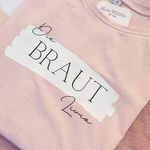 May include: A pink t-shirt with a white label that says "Mrs. Kiddo Est. 2023". The shirt has a white patch with the text "Die Braut Livia" in black.