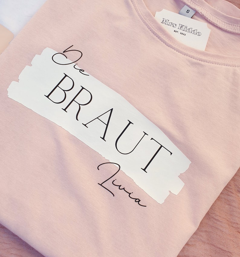 May include: A pink t-shirt with a white label that says "Mrs. Kiddo Est. 2023". The shirt has a white patch with the text "Die Braut Livia" in black.
