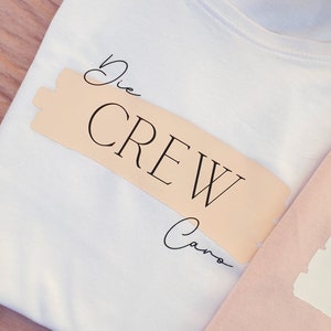 May include: White t-shirt with a peach-coloured label that says "Die Crew Caro" in black script.