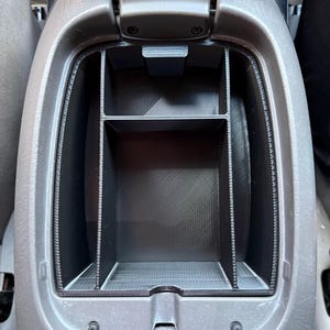 May include: A black plastic center console organizer insert with a grid-like structure featuring multiple compartments. The organizer is fitted inside a gray automotive center console storage bin, providing segmented storage space for small items.