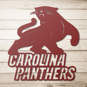 Puede incluir: Recorte met&aacute;lico granate de una pantera rugiendo. Las palabras "CAROLINA PANTHERS" est&aacute;n debajo. El recorte est&aacute; montado sobre un fondo de vetas de madera clara.