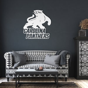 Puede incluir: Logotipo blanco de los Carolina Panthers, con un felino y el nombre del equipo, en una pared oscura. El logotipo est&aacute; encima de un sof&aacute; con un patr&oacute;n geom&eacute;trico en blanco y negro, una mesa de centro y una alfombra.
